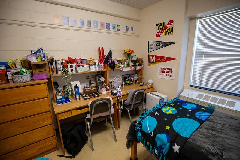 A desk setup in a residence hall room featuring two chairs, Maryland pennants, and a planet-print bed.