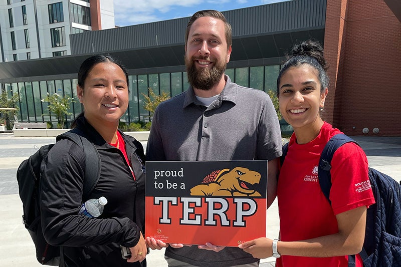 Three staff members holding a Proud Terp sign outside on Heritage Plaza.