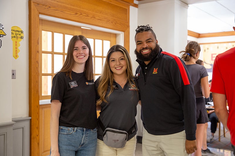 Three smiling staff members in UMD apparel posing together in a building hallway.