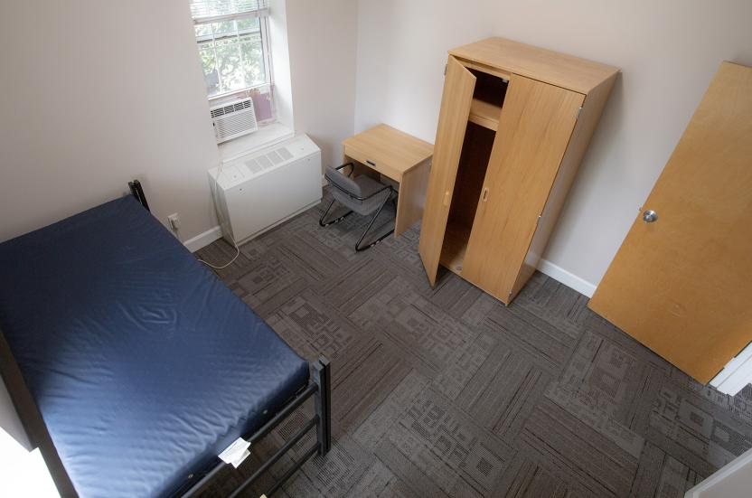 Aerial view of a single residence hall room with carpet flooring, a window, a twin bed, a desk with chair and wardrobe.