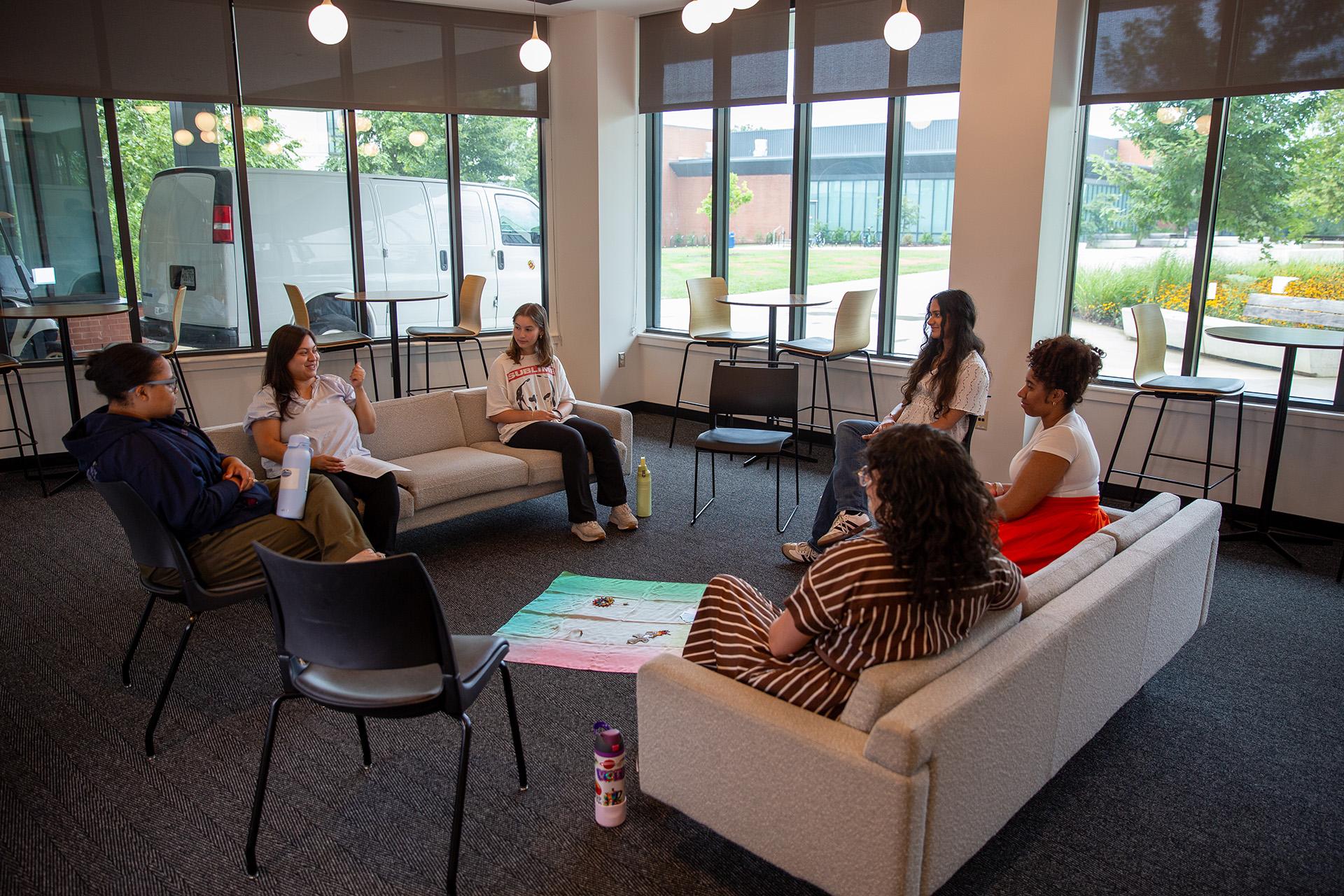 Six people seated in a circle on couches and chairs inside a bright lounge space, engaged in a group discussion.