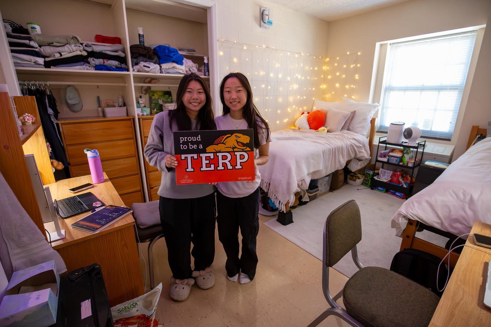 Two smiling students hold a “Proud to be a TERP” sign in a cozy dorm room.