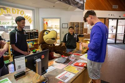 A person in a large brown turtle mascot costume accepts an ID card across a front desk while two smiling staff members stand behind the front desk.