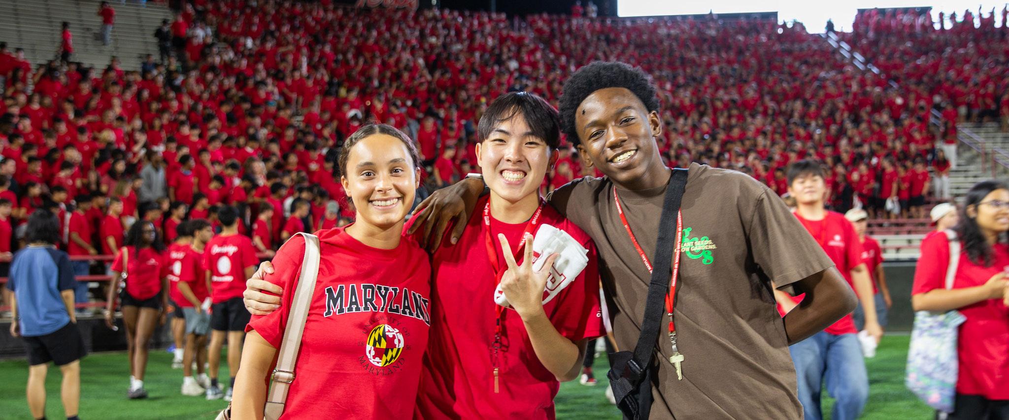 Three smiling students posing on a field with a crowded stadium in the background.