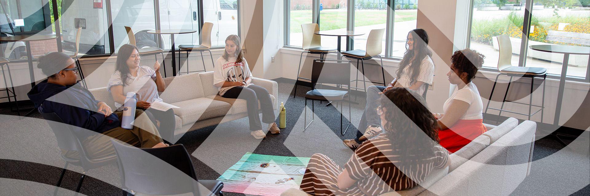 Six people seated in a circle on couches and chairs inside a bright lounge space, engaged in a group discussion.