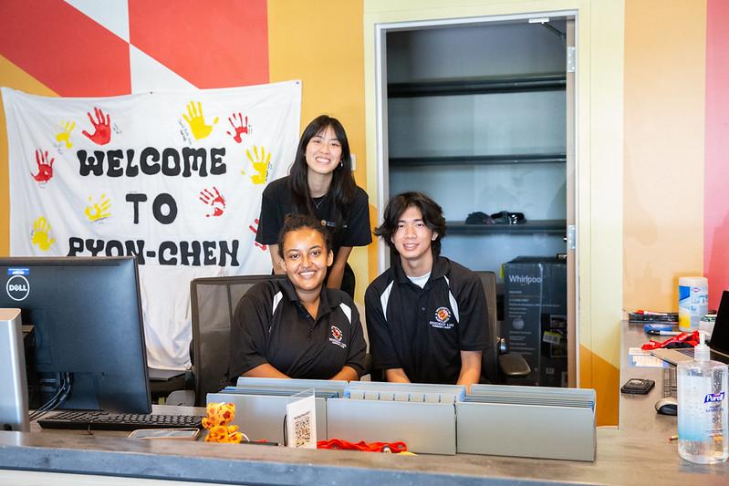 Three student staff members at a desk with a "Welcome to Pyon-Chen" banner.