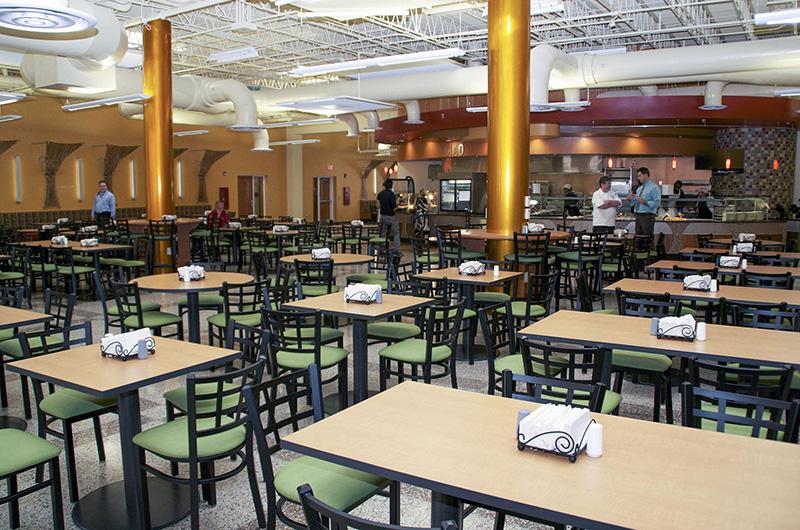 A spacious dining hall with many green-cushioned chairs, wooden tables, and large gold pillars.