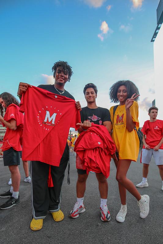Three students pose at SECU Stadium, one holding a red "Maryland" t-shirt, during the Big Show event.
