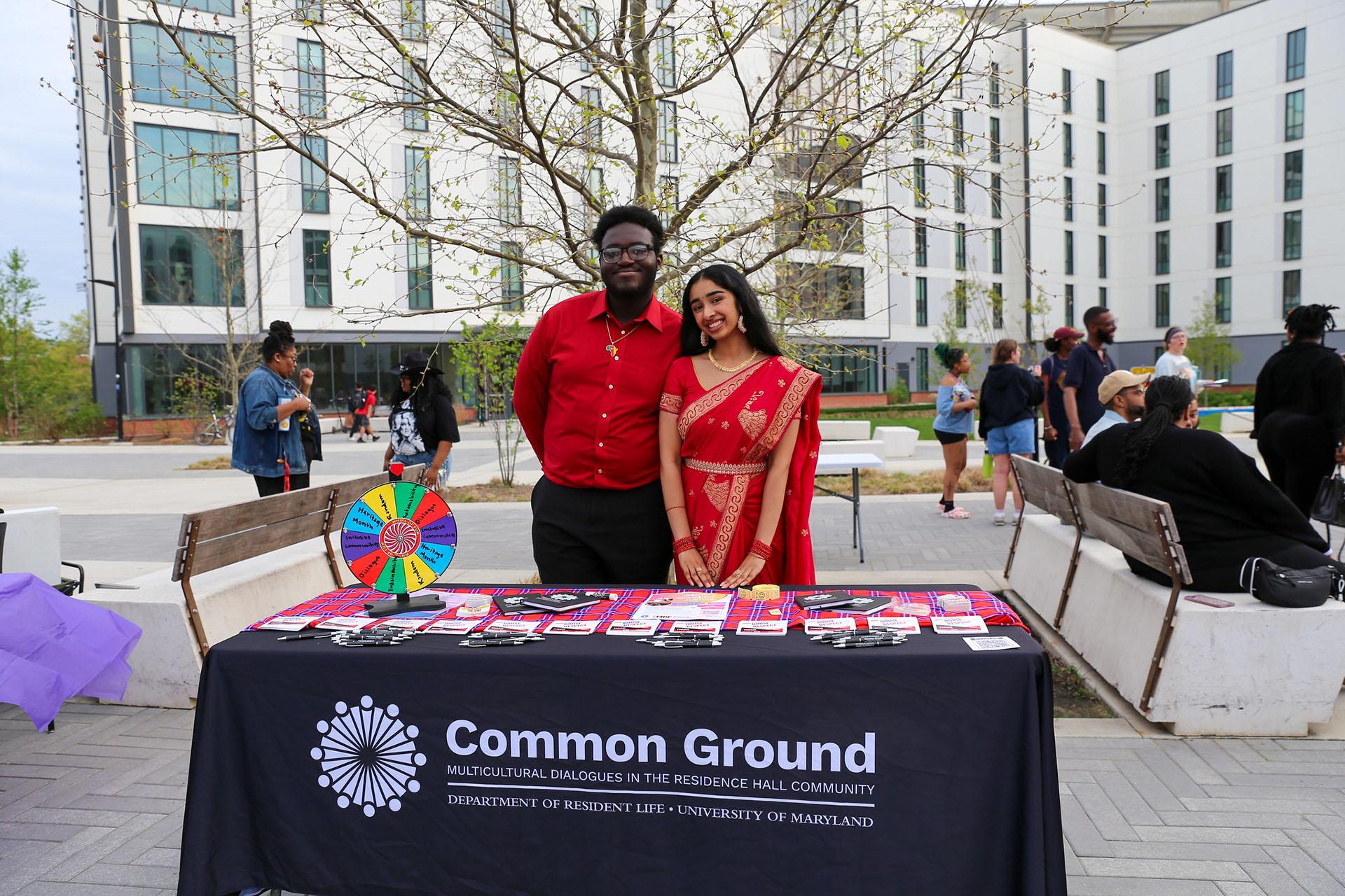 Two students pose behind a "Common Ground" table during the outdoor Heritage Fashion Show event.