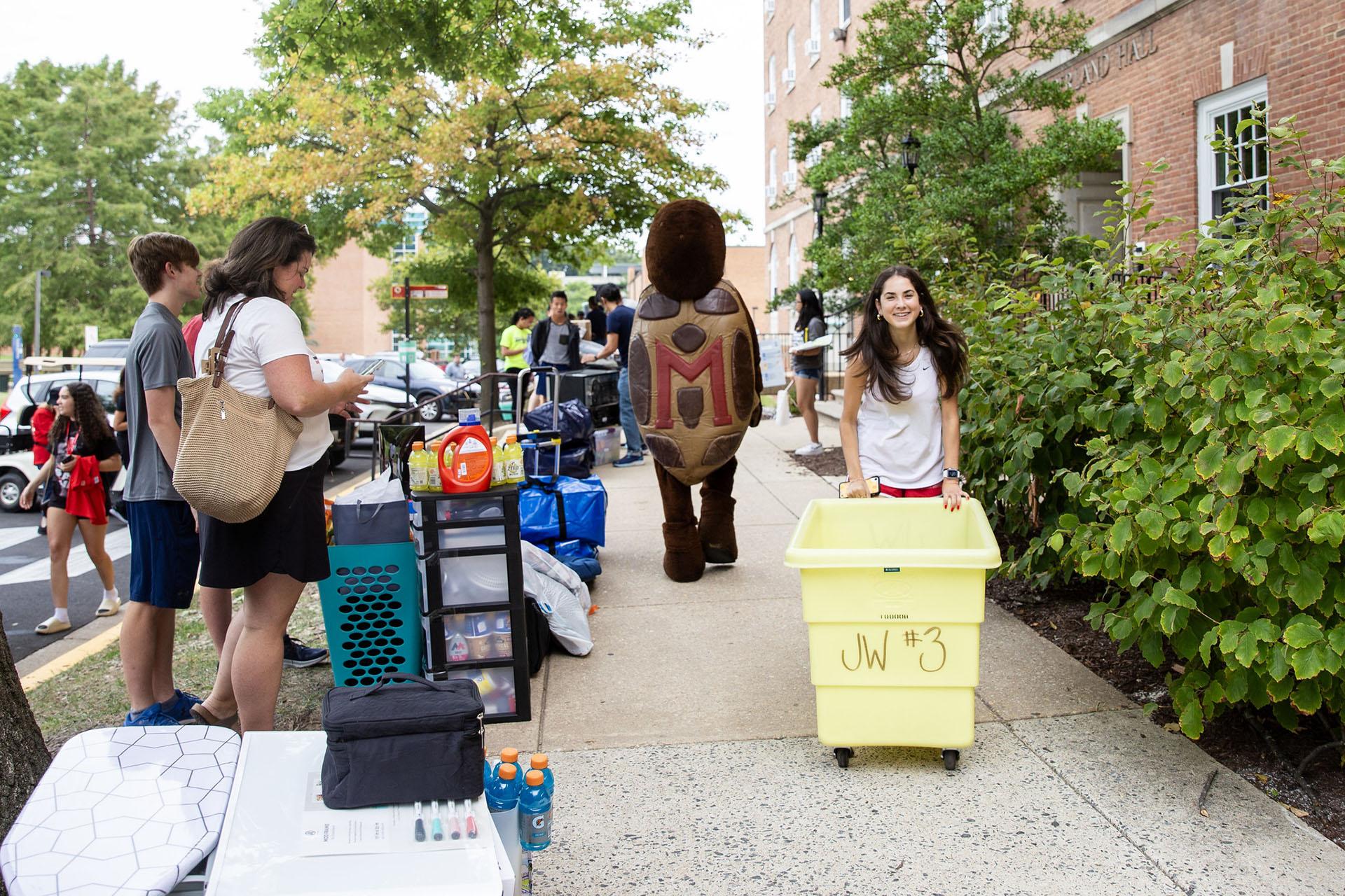 A smiling student pushes a large yellow moving cart on a sidewalk during campus move-in day with Testudo in the background.