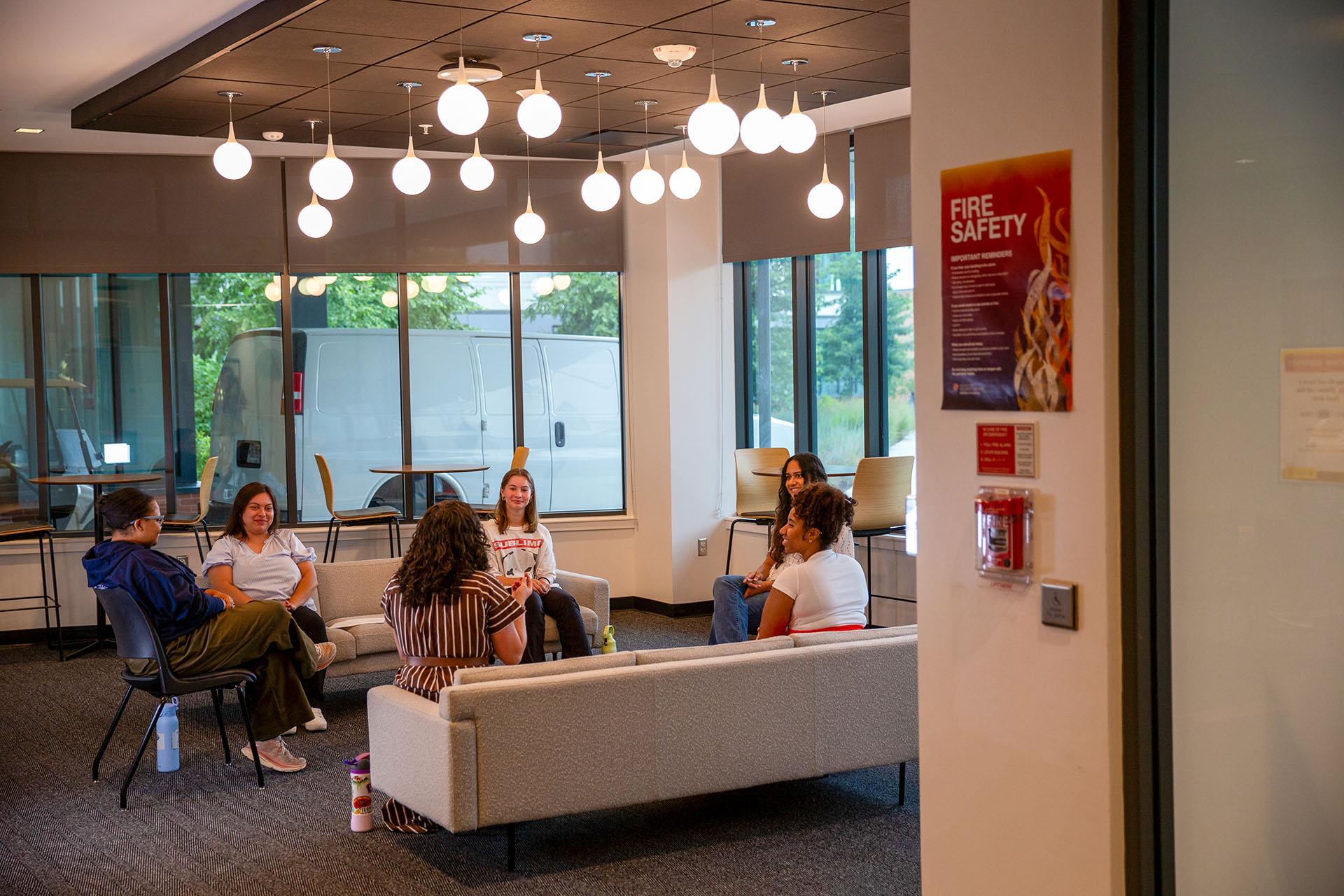 Five people sit in a circle on sofas in a modern lounge area beneath globe pendant lights.