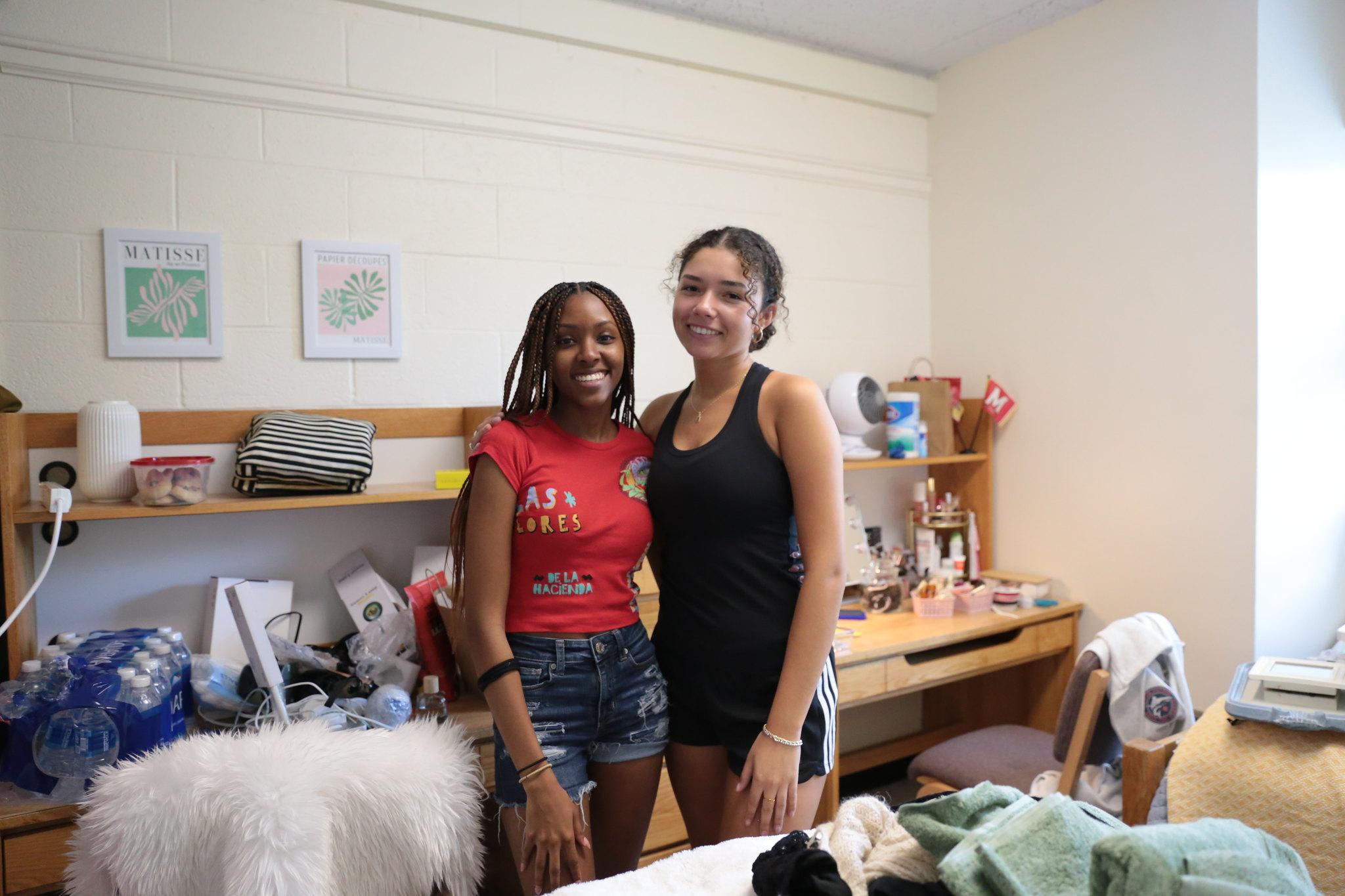 Two female students smiling and posing in a dorm room filled with boxes and move-in supplies.
