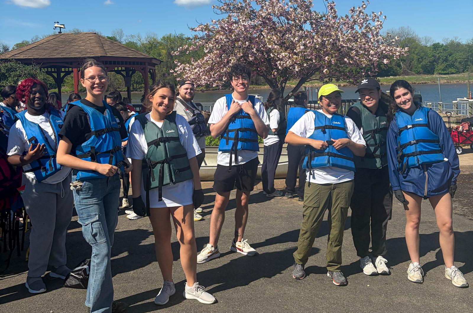 Smiling volunteers in life jackets pose near a waterfront and flowering tree.
