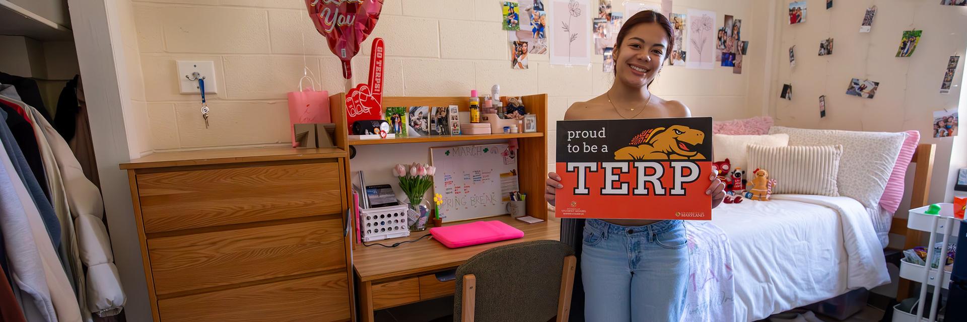 A student in her residence hall room, with a bed to her right and closet to her left, holding a Proud to be a Terp sign.