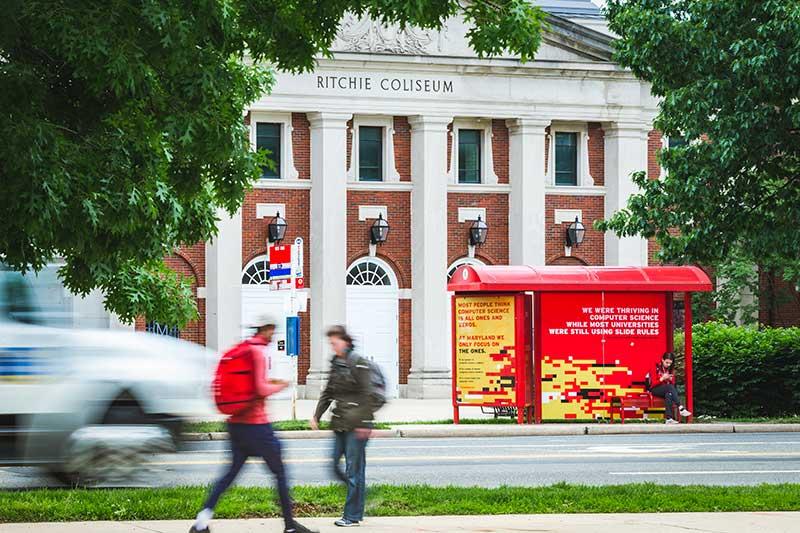 Red bus shelter on a busy street in front of the white columns of Ritchie Coliseum.
