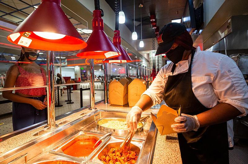 A dining services worker in a mask and apron serves food into a takeout container at a buffet.