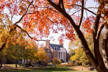 A brick campus building with white columns is framed by vibrant red and orange autumn leaves.